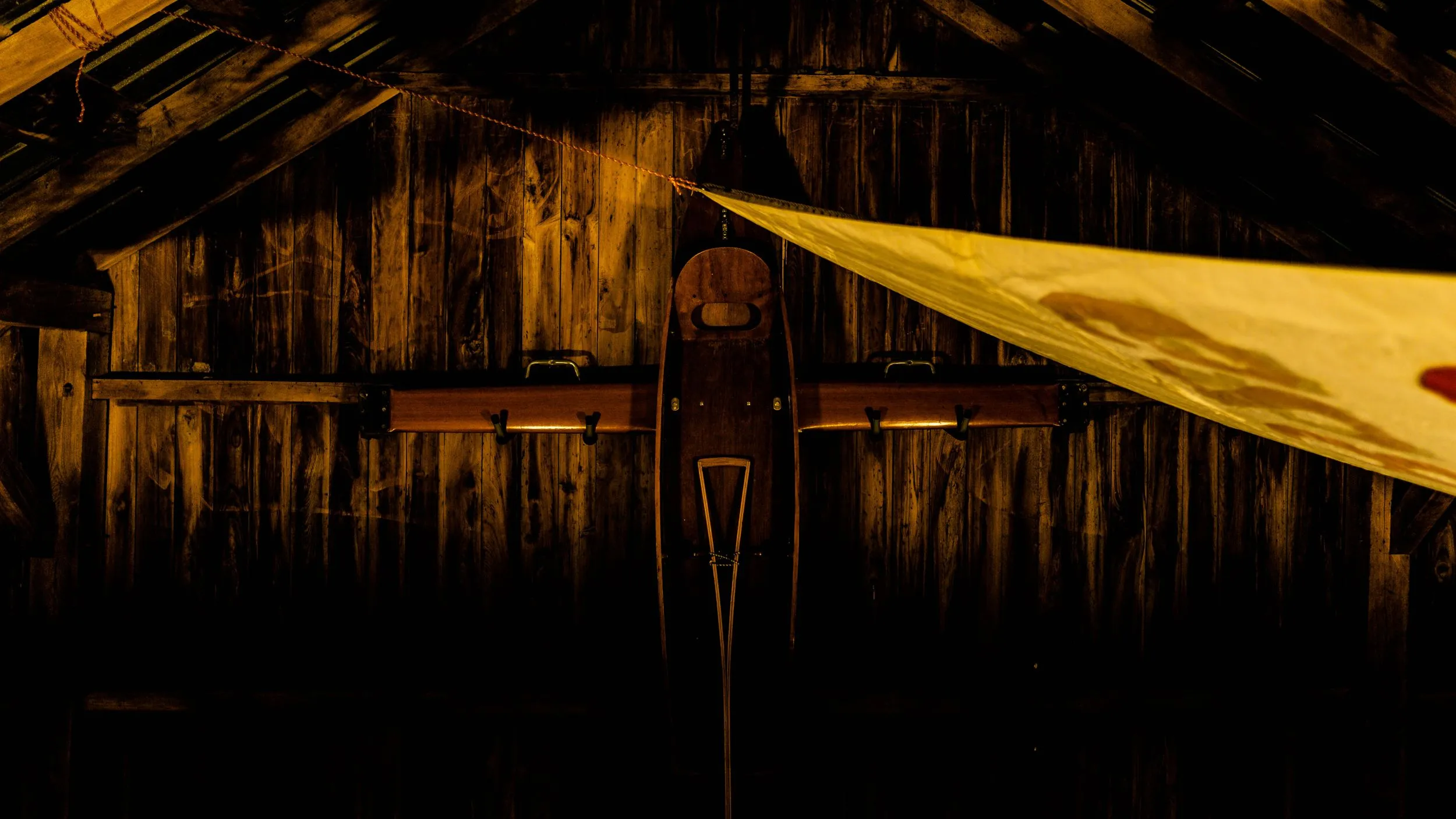 Wooden rowing shell and oars mounted on a dim barn wall