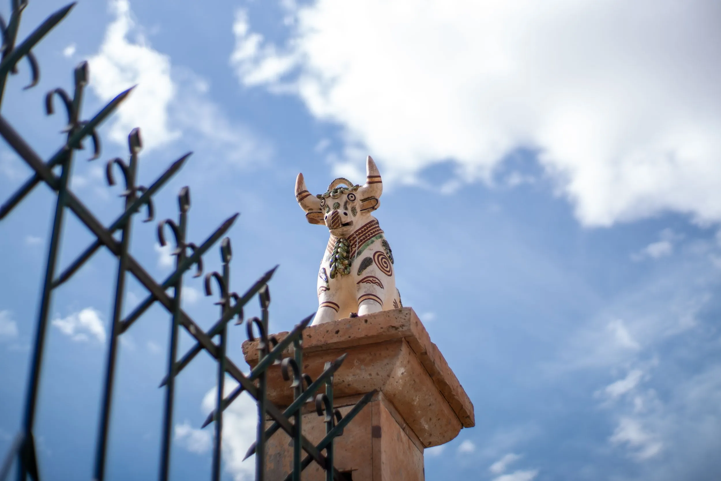Painted ceramic bull figure atop a stone pillar beside an iron fence against sky